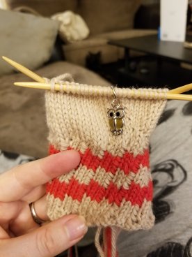 Knitted mitten with cream colored background with red diagonal stripes, shown on knitting needles.